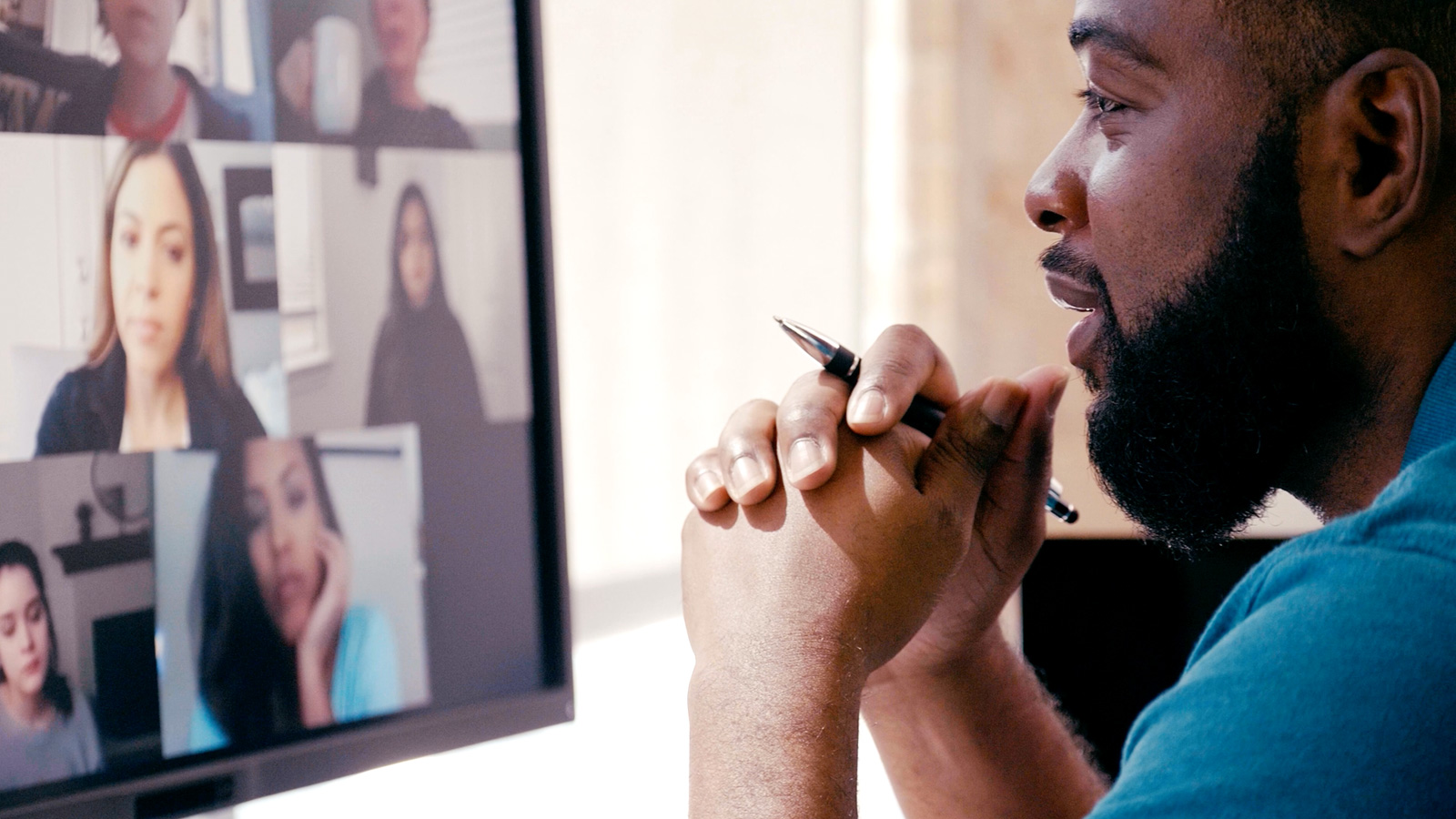 A black man sitting, holding a pen, looking at a computer monitor showing a grid of coworkers' faces