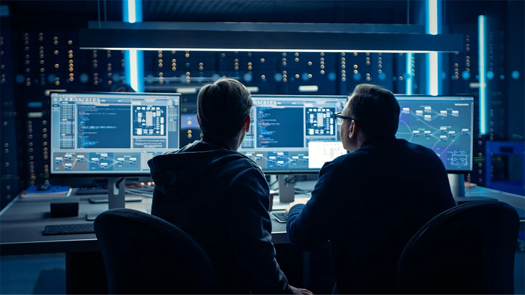 Two men sitting at a desk looking at monitors in front of a server room