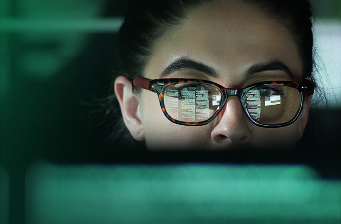 Close-up of a person with dark hair tied back, wearing thin-framed glasses, against a green-toned background.