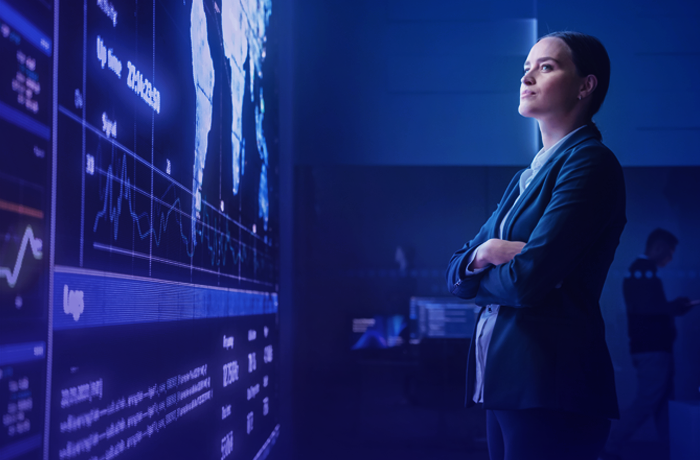 Person in a suit standing with arms crossed, looking at large digital screens displaying maps and data charts in a blue-lit room.
