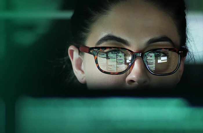 Close-up of a person with dark hair tied back, wearing thin-framed glasses, against a green-toned background.