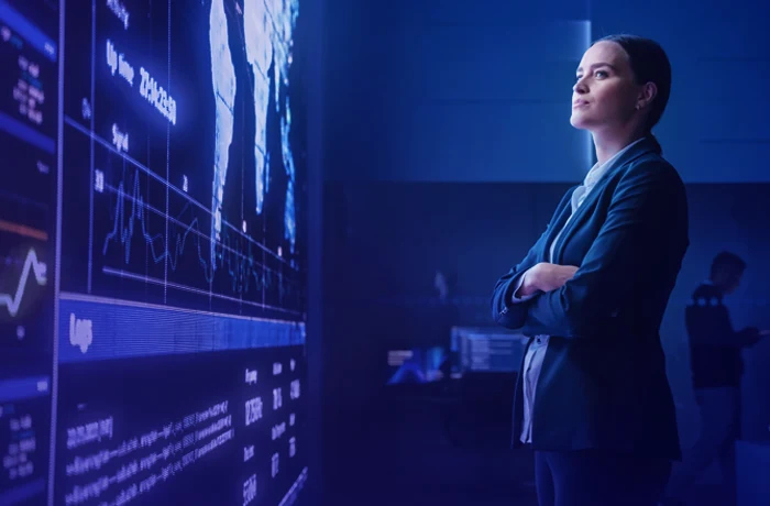 Person in a suit standing with arms crossed, looking at large digital screens displaying maps and data charts in a blue-lit room.