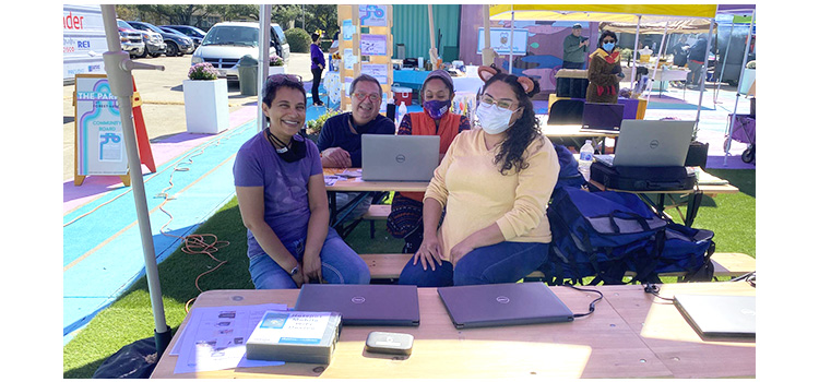 People sitting at desks with laptops under an outdoor tent, smiling