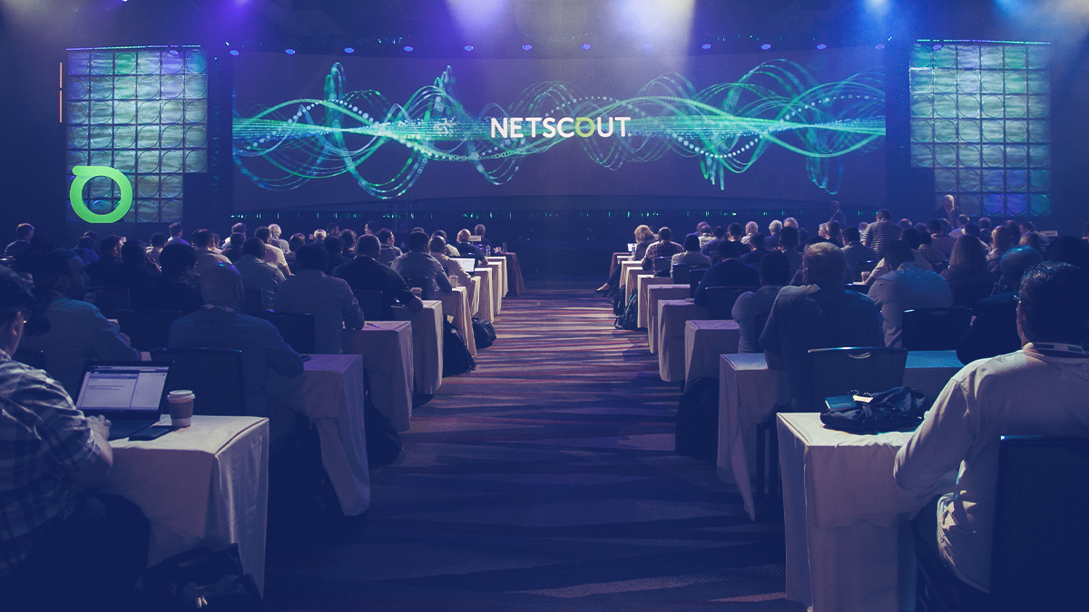 A large conference room full of attendees sitting at tables, with a large dais and screen with the NETSCOUT logo in front of them.