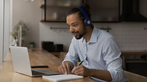 Man at desk looking at laptop with headphones and notepad