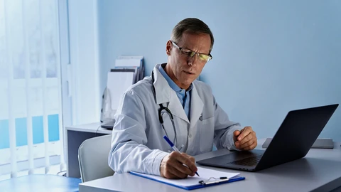 Male doctor in white lab coat sitting at desk with laptop and clipboard