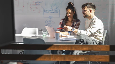 Man and Woman sitting at desk looking over laptop with whiteboard in background