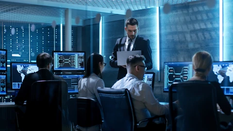 Four people sitting behind monitors in data center with One person standing holding a laptop