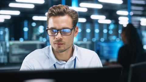 Young man wearing glass sitting in datacenter looking at monitors
