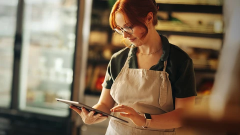 A woman in a white apron holding a tablet and what appears to be reviewing an order.