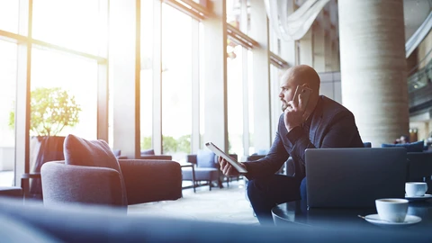 Gentlemen looking out windows on cell with tablet in his hand.