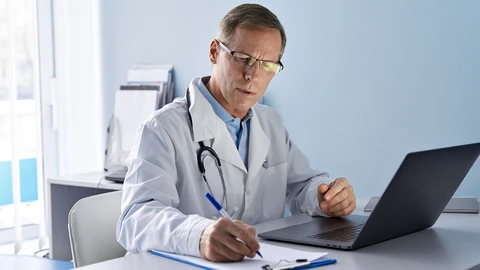 Doctor using application on laptop while reviewing information on clipboard
