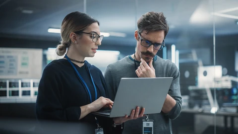1 person holding a laptop while another one stands beside while reviewing something being presented on laptop. 