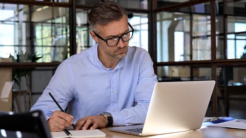 Businessman with glasses looking at laptop writing on tablet.