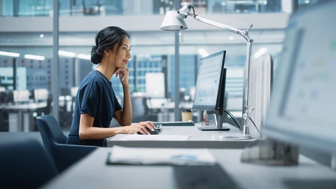 Woman in front of computer with datacenter to the left of her.