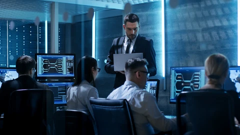 Man in suit with laptop in front of people behind desktops