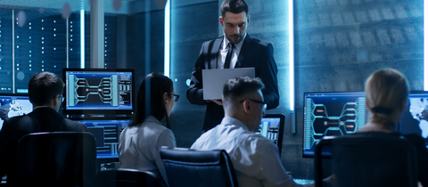 People sitting at desk looking at monitors behind glass wall in a datacenter.
