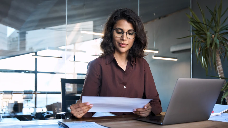 Woman reviewing KPI's on a sheet of paper and laptop to her left